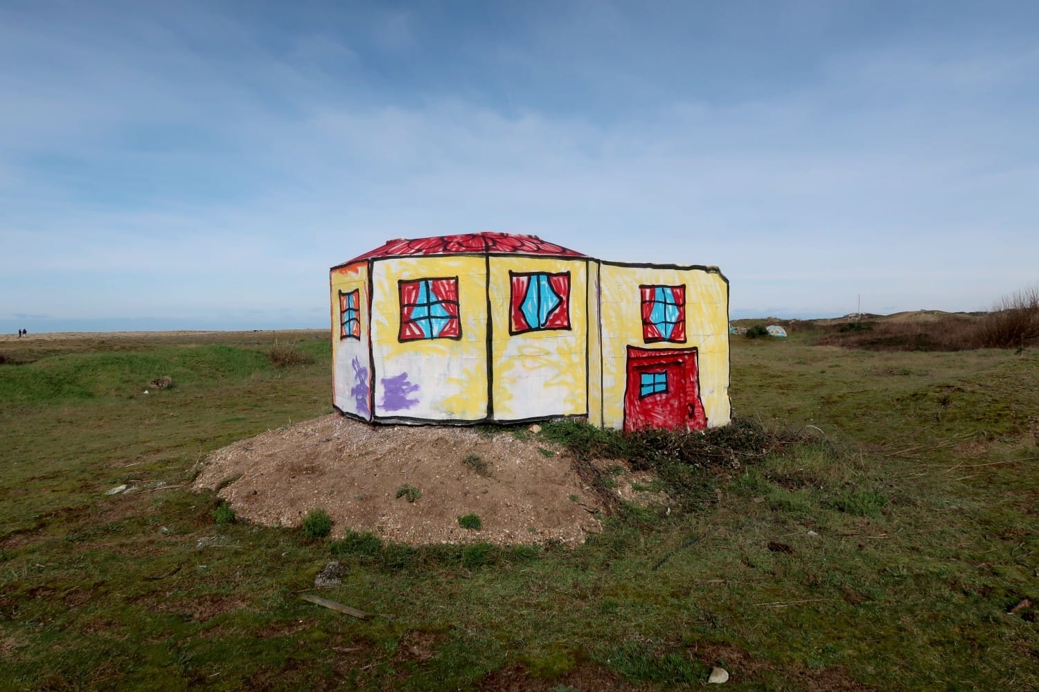 Betonnen bunker in een graslandschap beschilderd als een geel huisje met een rood dak, ramen en een rode deur in kindertekenstijl